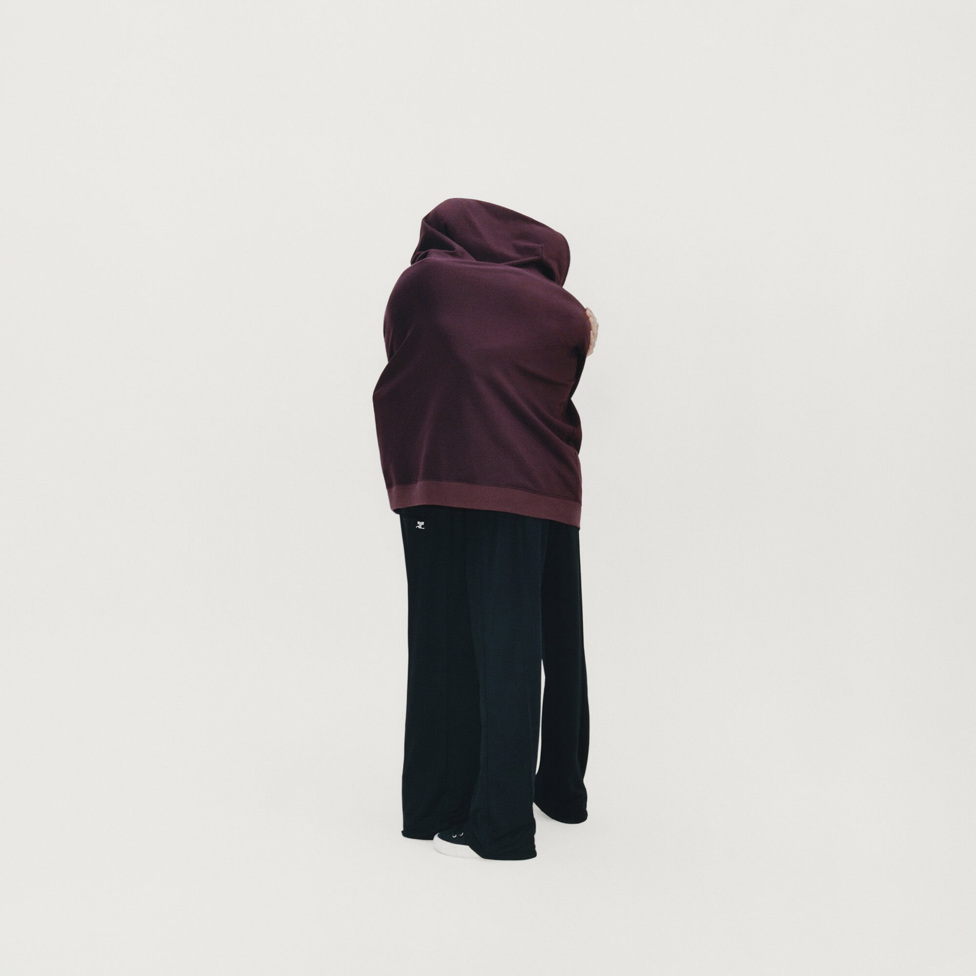 Two people standing closely together wrapped in a large burgundy hooded garment against a plain white studio background.