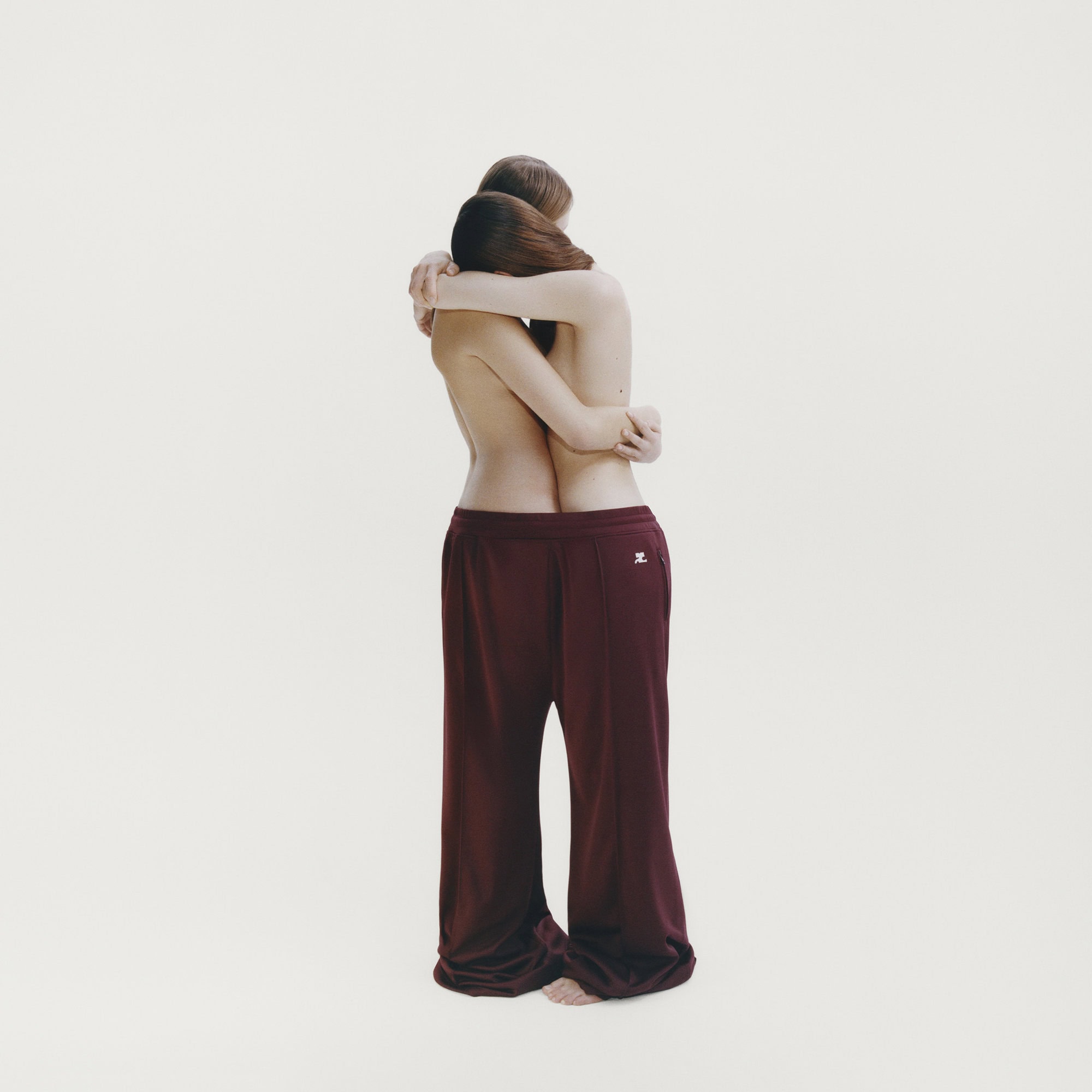Two people embracing while wearing a single oversized pair of burgundy trousers against a white background