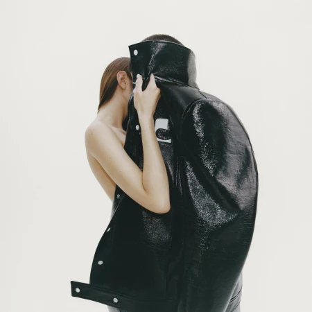 Model holding a glossy black vinyl jacket in front of their face against a plain white background