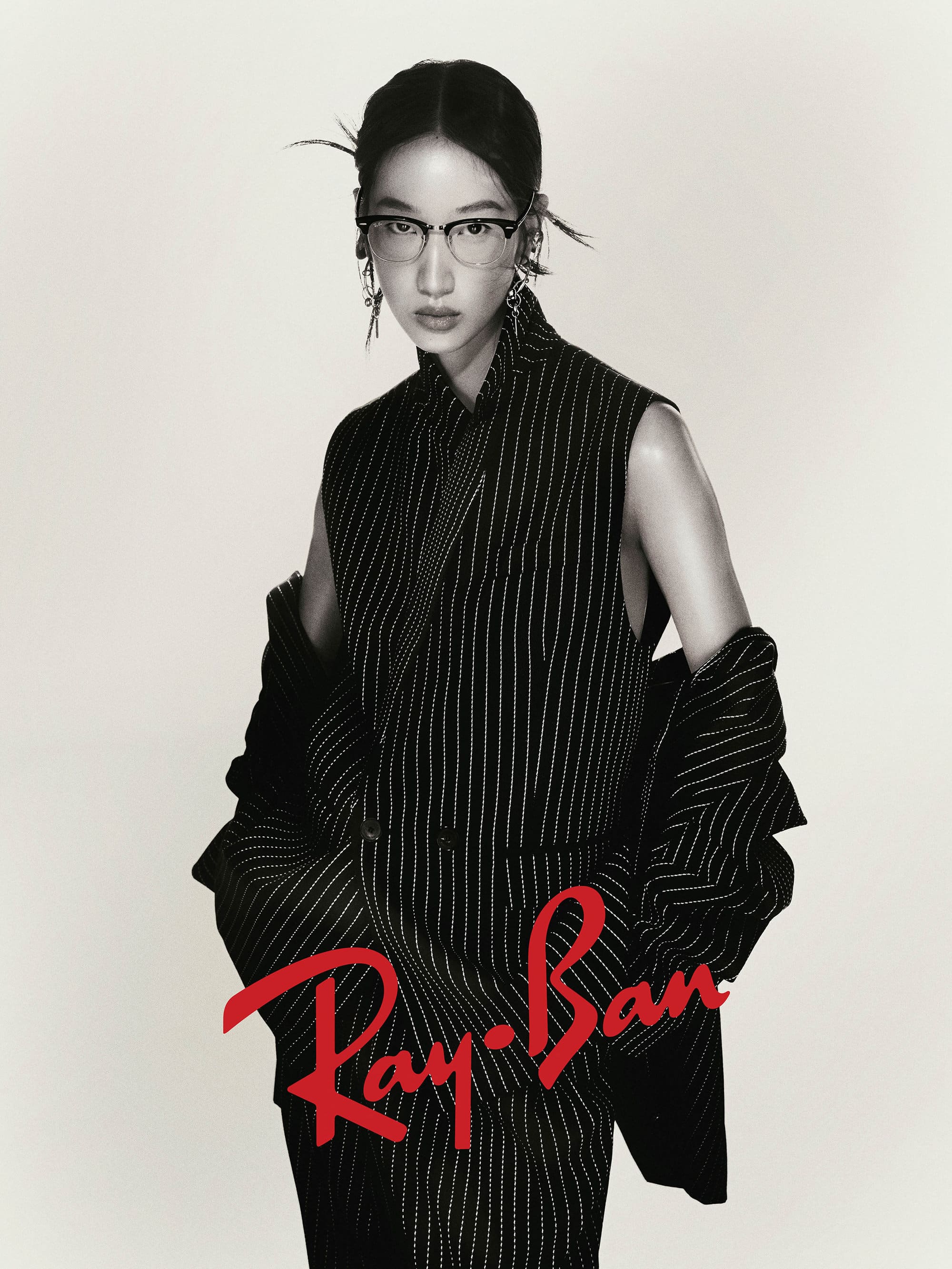 Model wearing black eyeglasses and a pinstripe sleeveless top with a draped jacket against a neutral backdrop