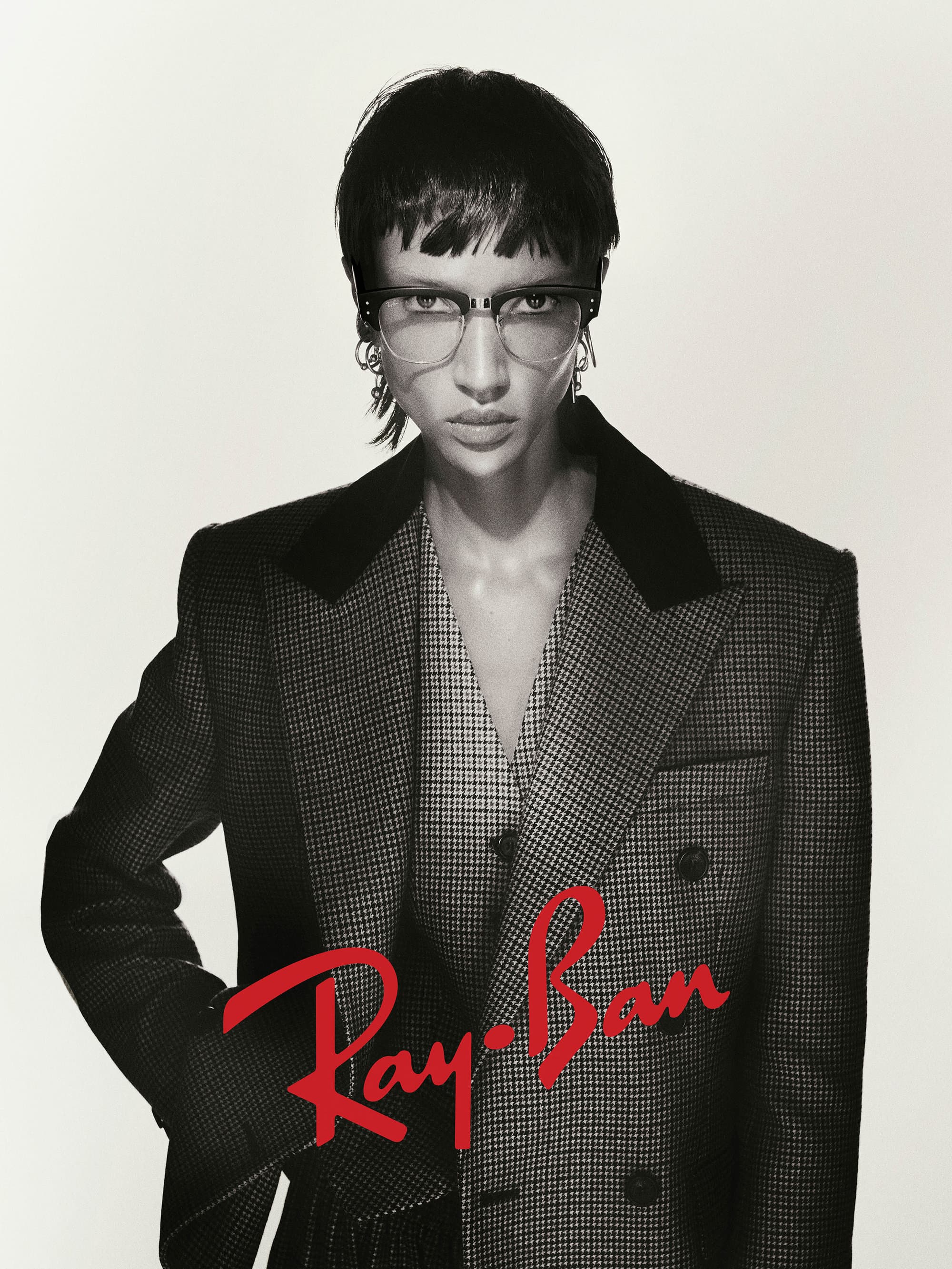Model wearing dark glasses and a patterned blazer against a neutral backdrop for the Ray-Ban 2025 campaign.