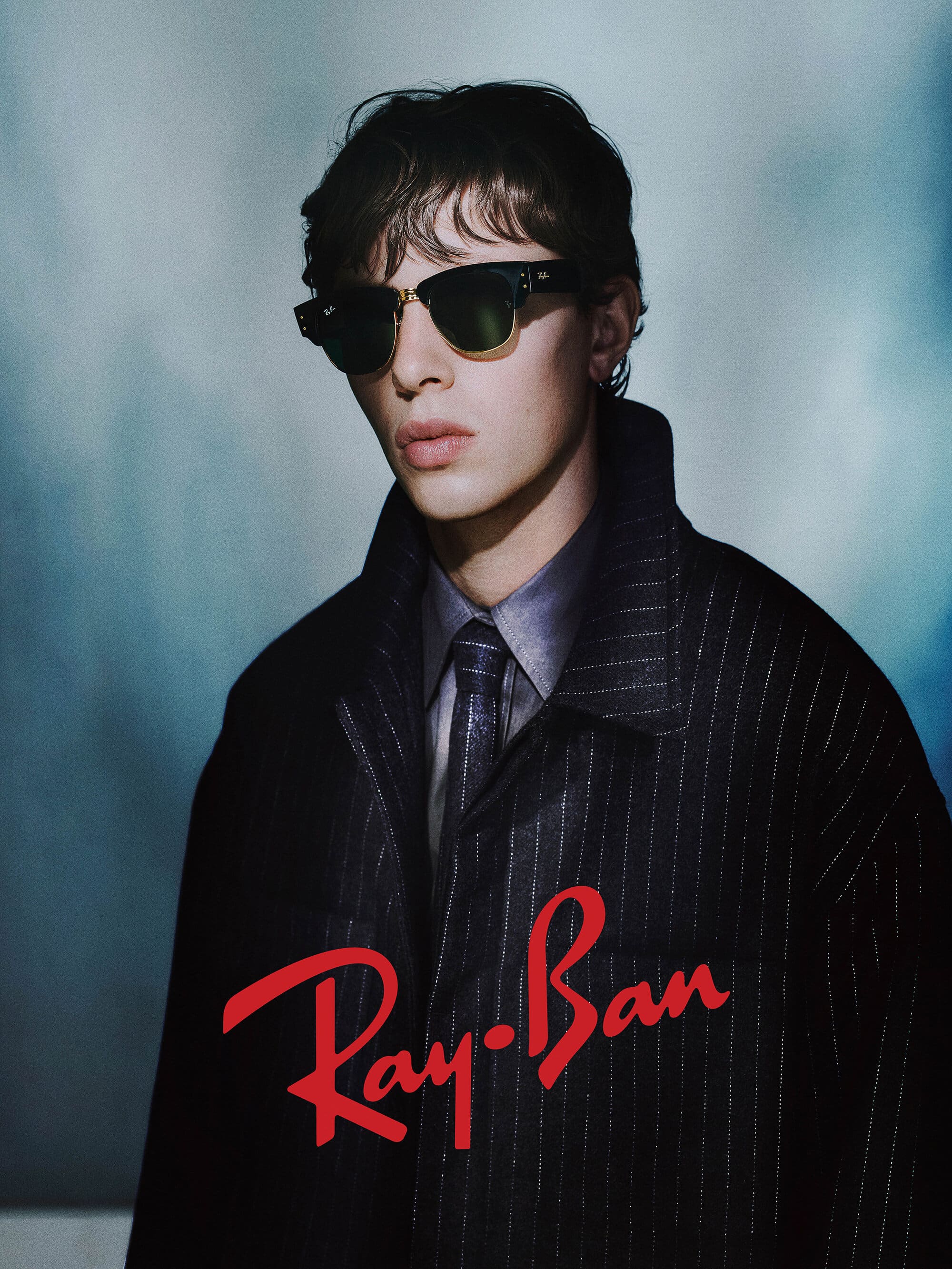 Young man wearing sunglasses and a pinstripe coat against an atmospheric blue background for the Ray-Ban 2025 campaign.