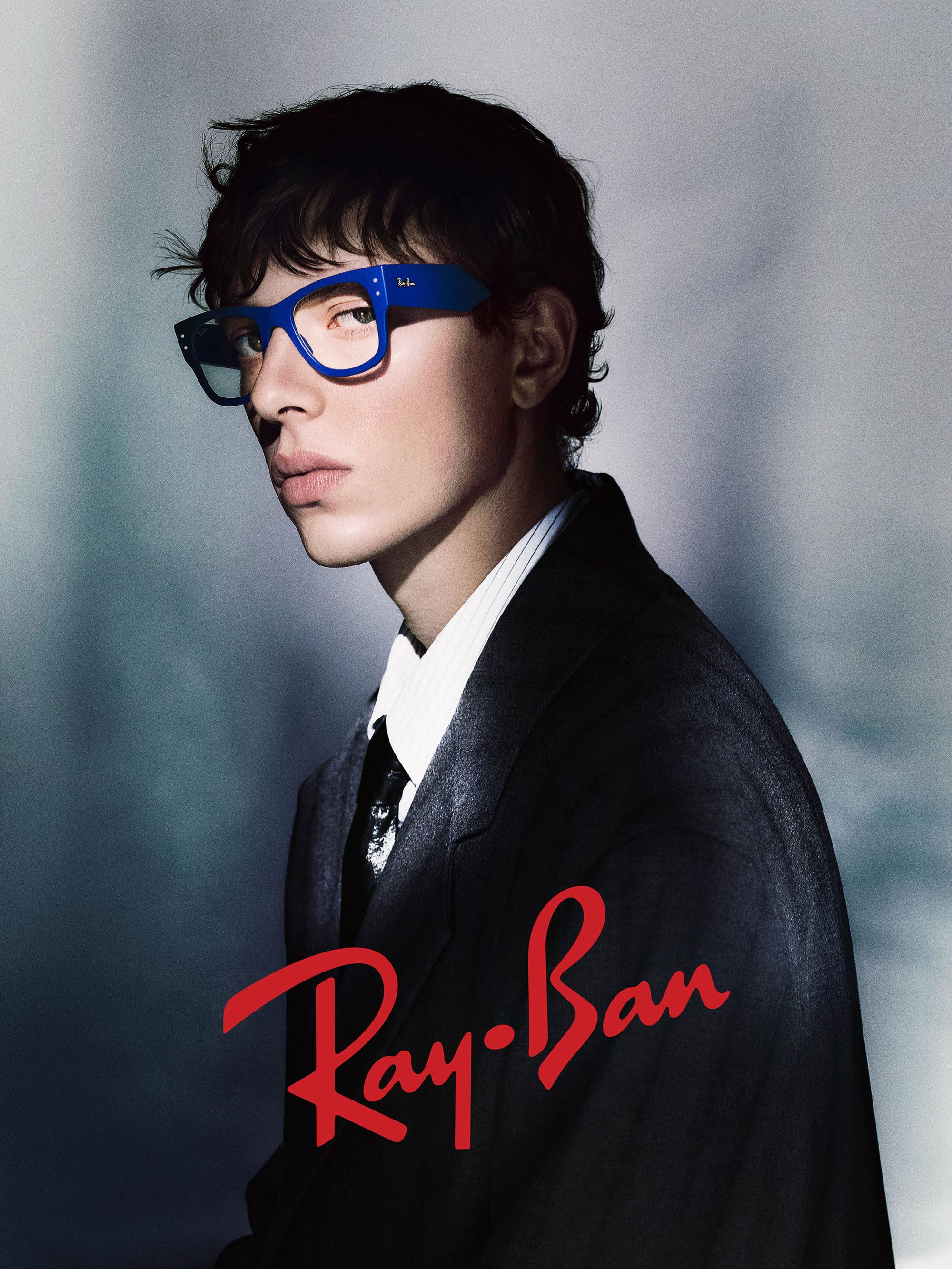 Young man wearing blue glasses and a black suit jacket against a grey seamless background for Ray-Ban The Icons ad campaign