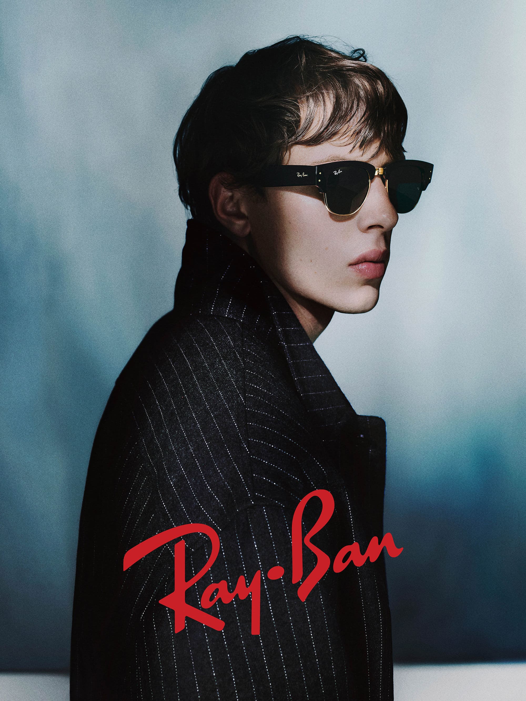 Close-up profile of a male model wearing dark sunglasses and a dark pinstripe coat against a muted background
