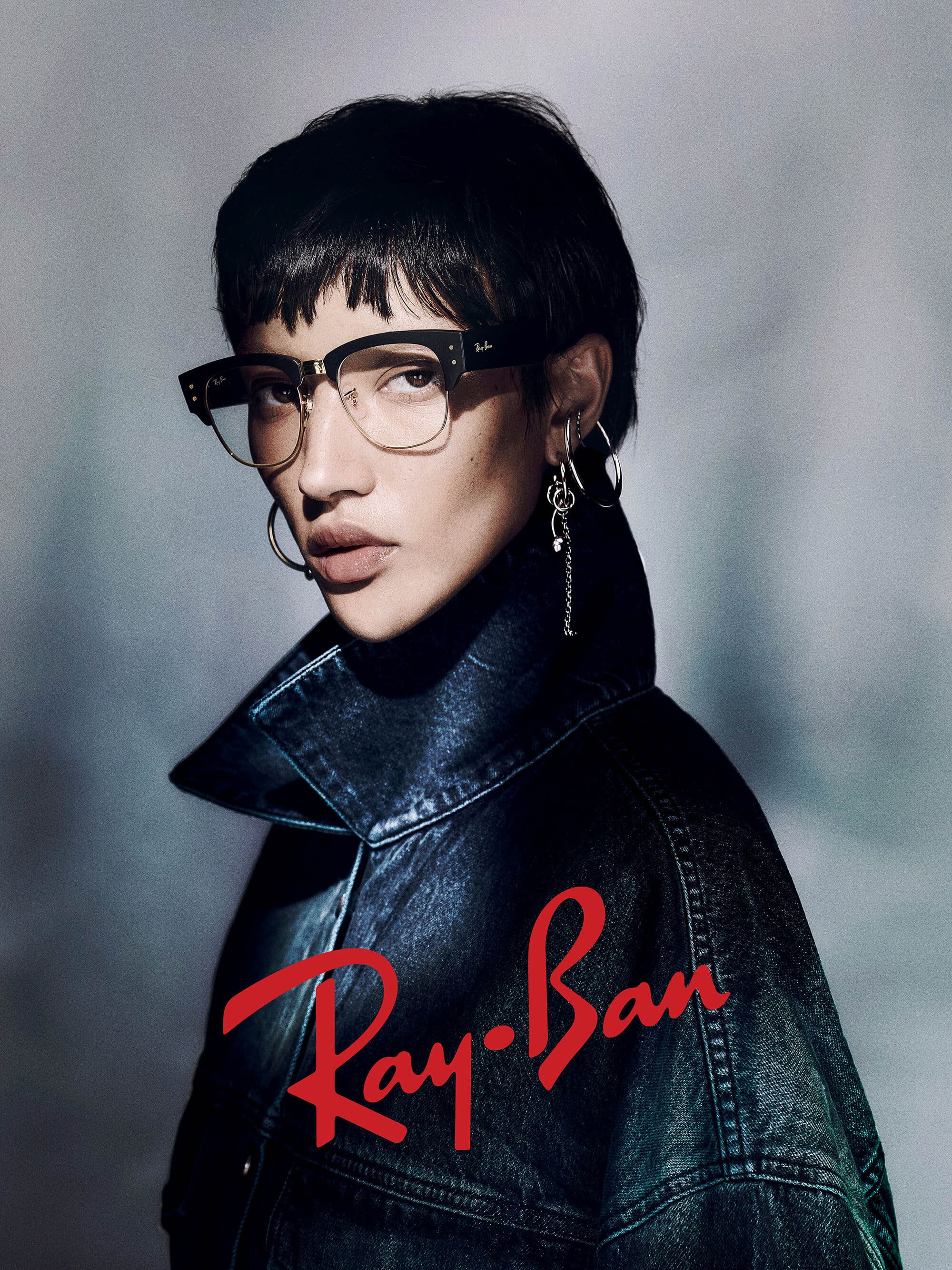 Model wearing dark framed glasses and a dark denim jacket against a textured grey background with red Ray-Ban logo
