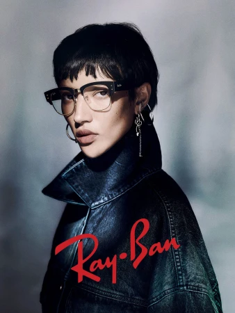 Model wearing dark framed glasses and a dark denim jacket against a textured grey background with red Ray-Ban logo