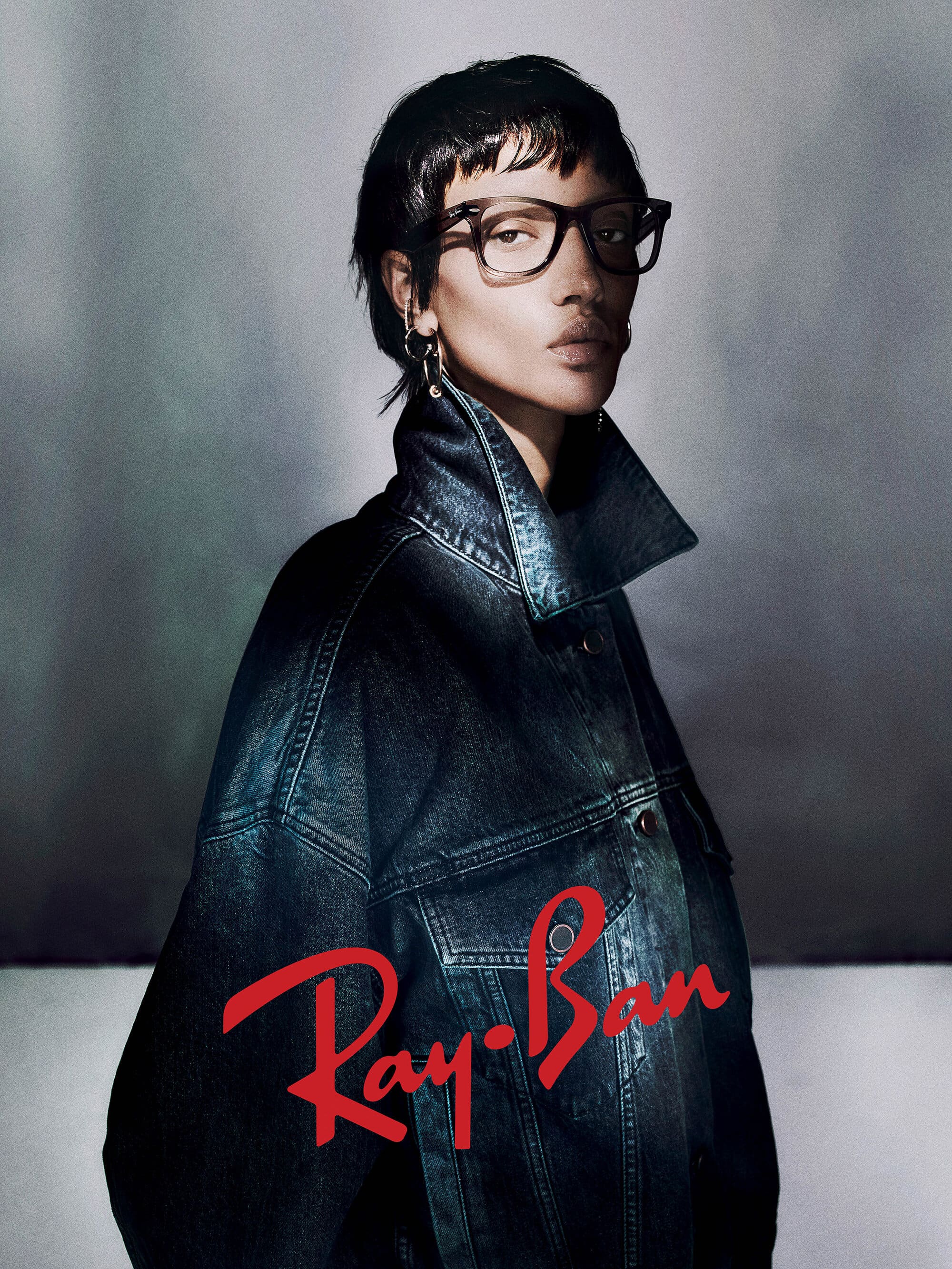 Model wearing dark eyeglasses and an oversized denim jacket against a textured grey background with Ray-Ban logo