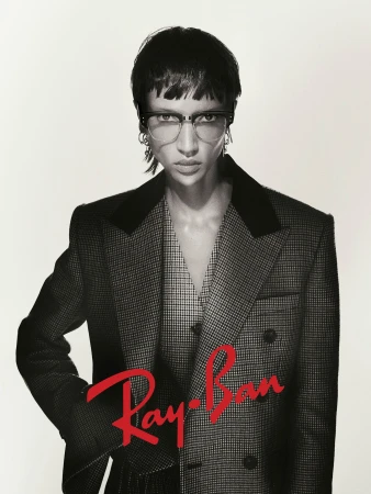 Model wearing dark glasses and a patterned blazer against a neutral backdrop for the Ray-Ban 2025 campaign.