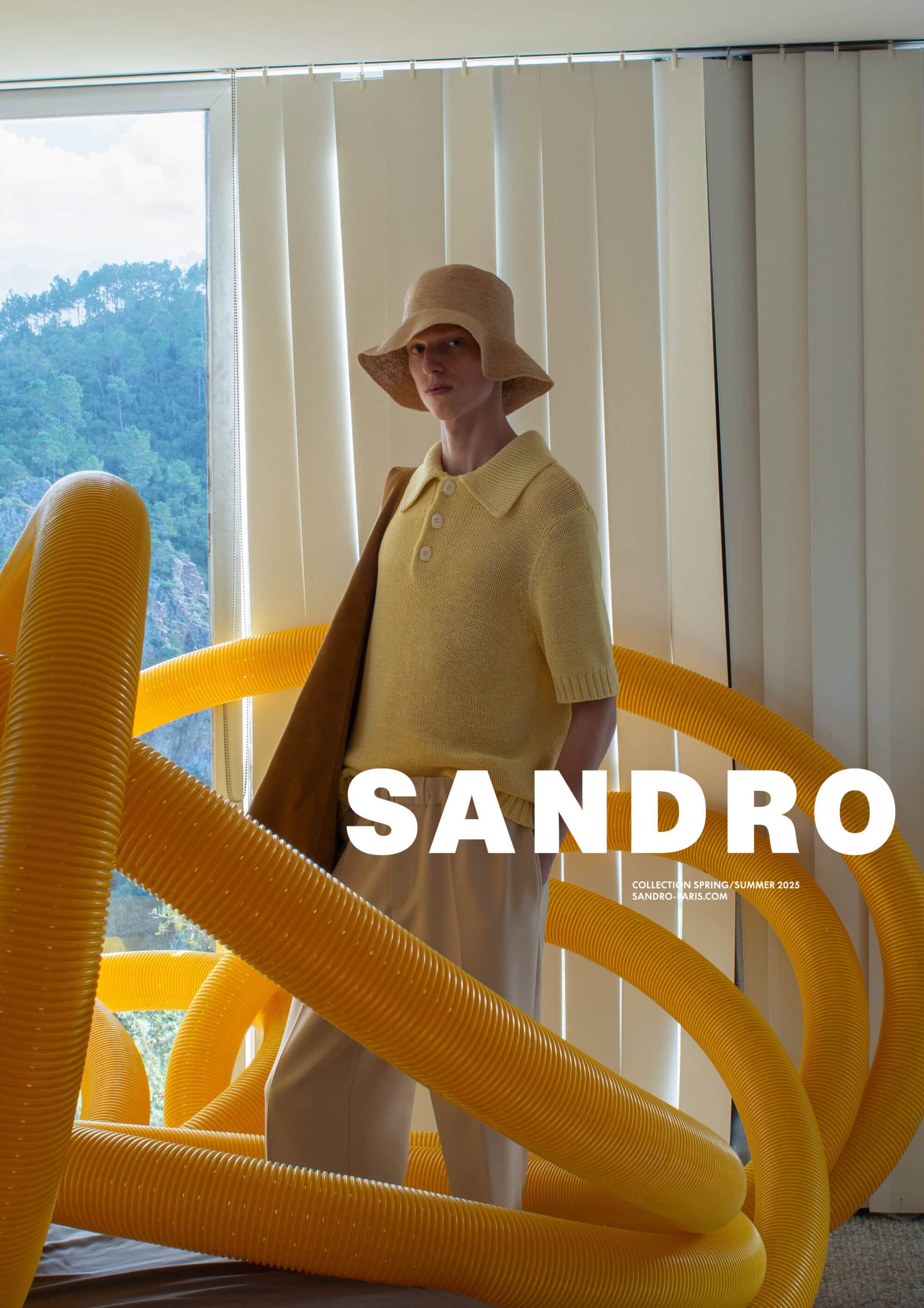 Model wearing a yellow polo shirt and bucket hat standing behind large yellow tubes in an indoor setting