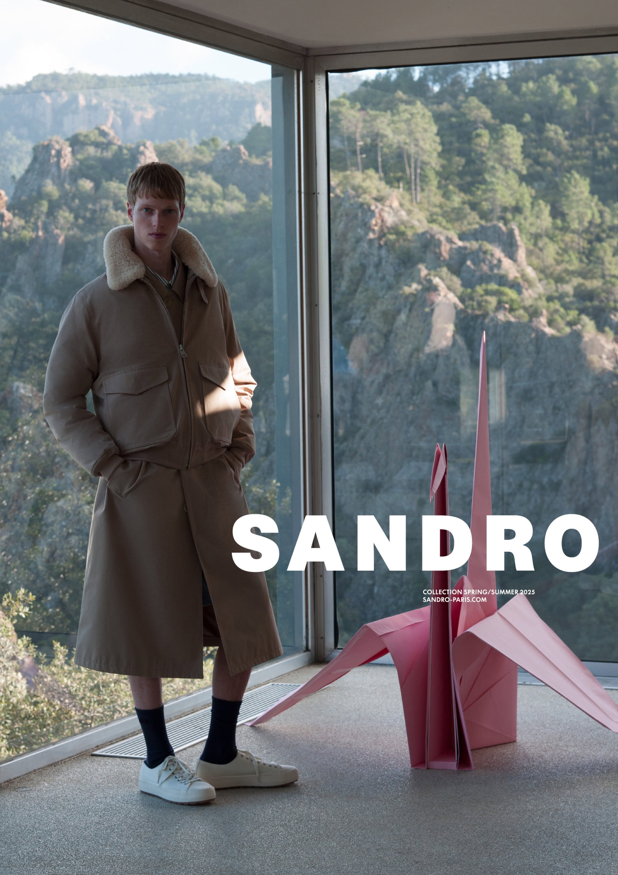 Male model standing indoors near a large window overlooking a mountainous landscape, next to a pink paper origami crane.