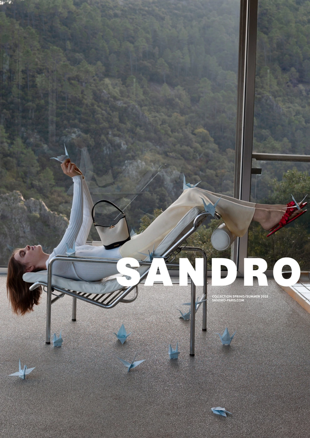 Model lying on a modern tubular steel lounge chair next to large glass windows overlooking a forest.
