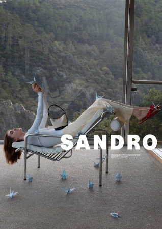 Model lying on a modern tubular steel lounge chair next to large glass windows overlooking a forest.