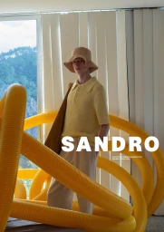 Model wearing a yellow polo shirt and bucket hat standing behind large yellow tubes in an indoor setting