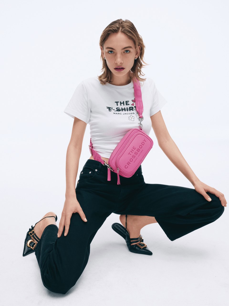 Model crouching against a white background wearing a white t-shirt, dark jeans, and a pink crossbody bag.