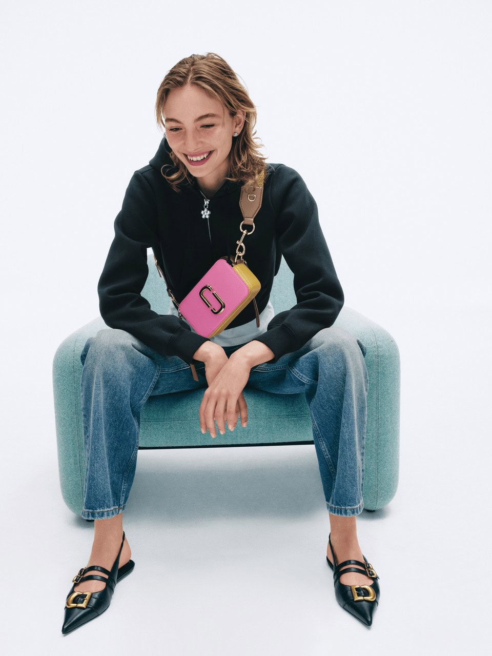 Smiling young woman seated in an armchair wearing a black hoodie, blue jeans, and slingback flats, carrying a pink and yellow crossbody bag.