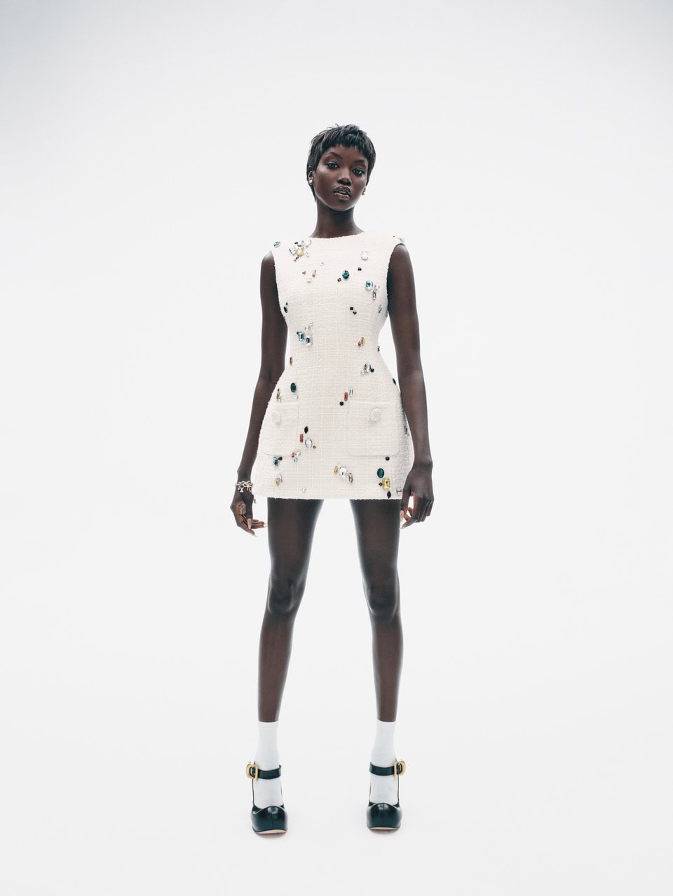 Anok Yai wearing an embellished white minidress and black Mary Jane shoes against a white seamless background.