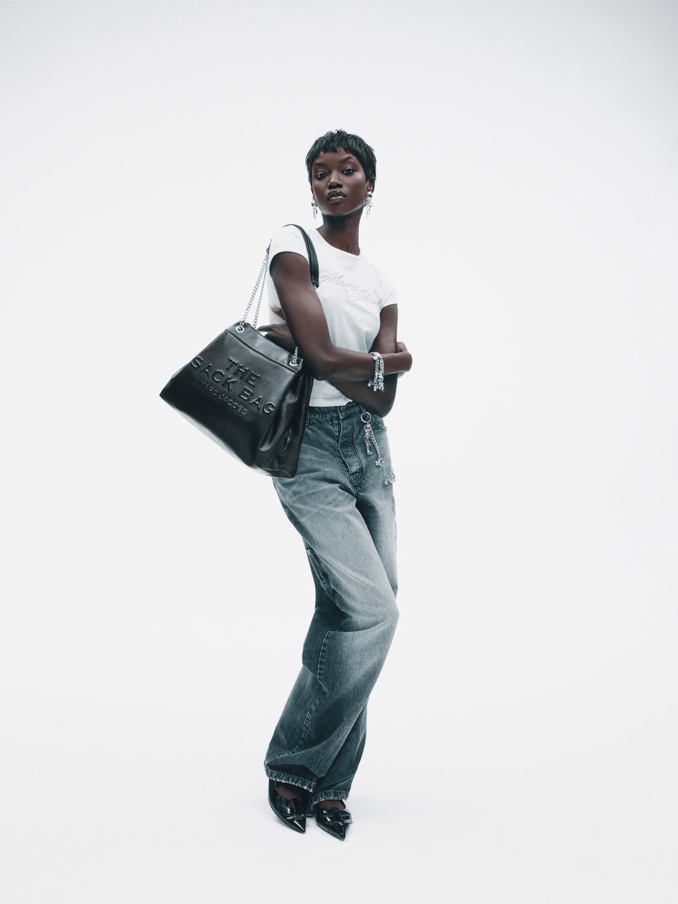 Anok Yai wearing a white t-shirt and grey jeans carrying a black leather handbag against a plain white background for Marc Jacobs.