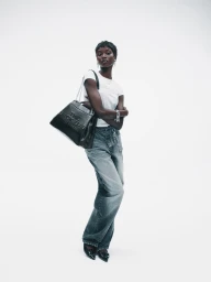 Anok Yai wearing a white t-shirt and grey jeans carrying a black leather handbag against a plain white background for Marc Jacobs.