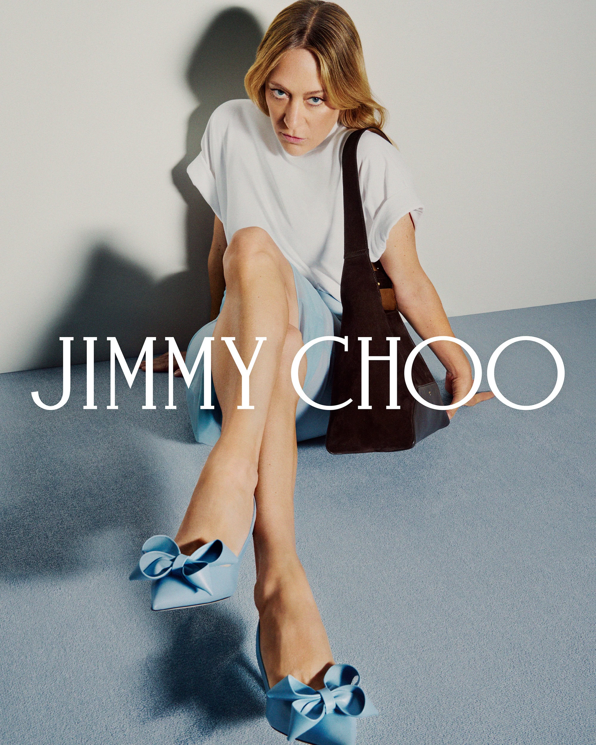 Chloe Sevigny seated on a blue floor wearing blue bow heels and a white top for Jimmy Choo Spring 2025 ad campaign.