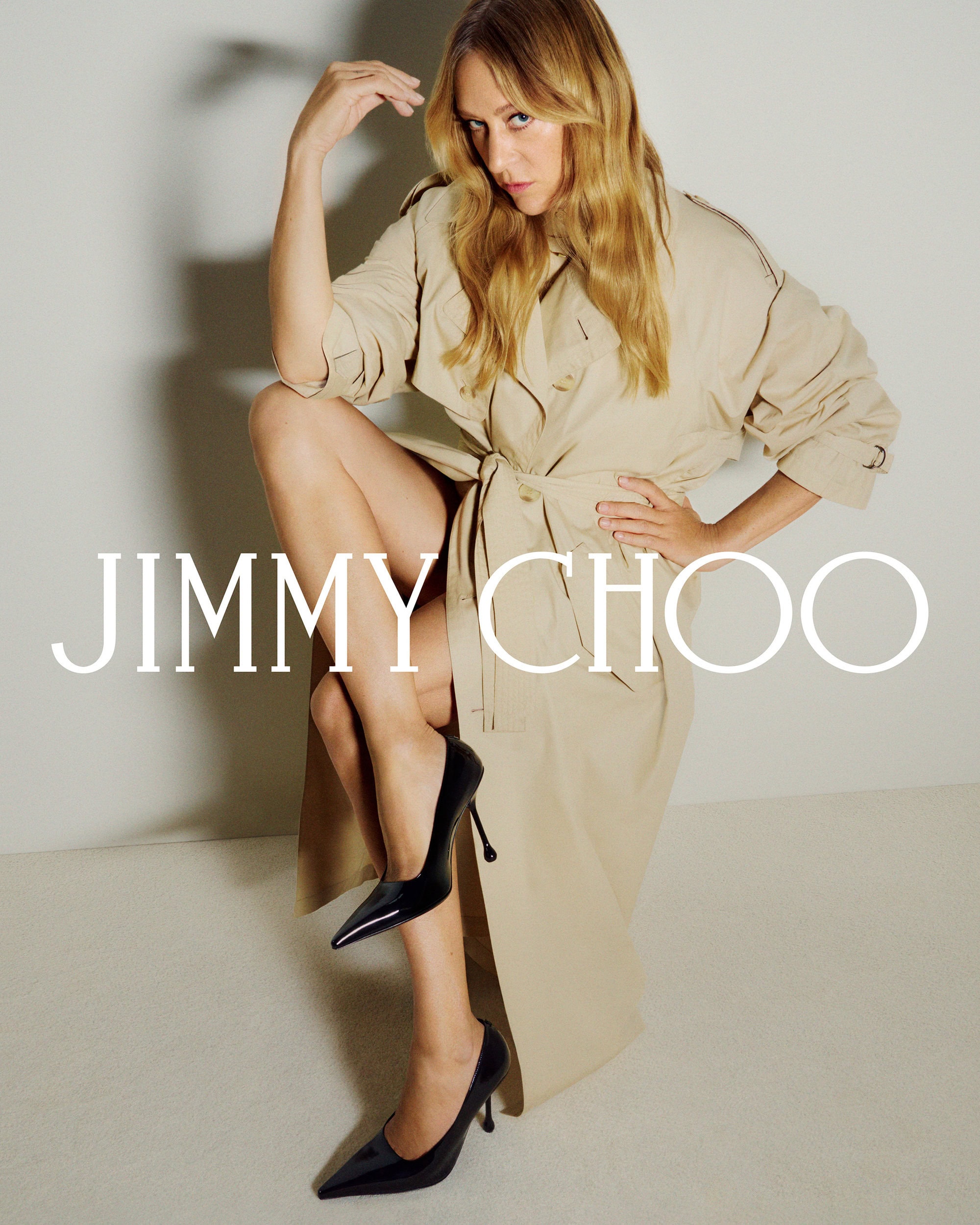 Chloe Sevigny wearing a beige belted trench coat and black heels against a light background for Jimmy Choo