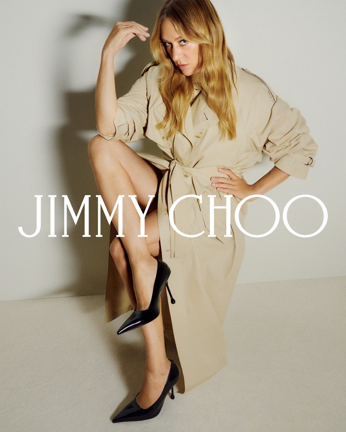 Chloe Sevigny wearing a beige belted trench coat and black heels against a light background for Jimmy Choo