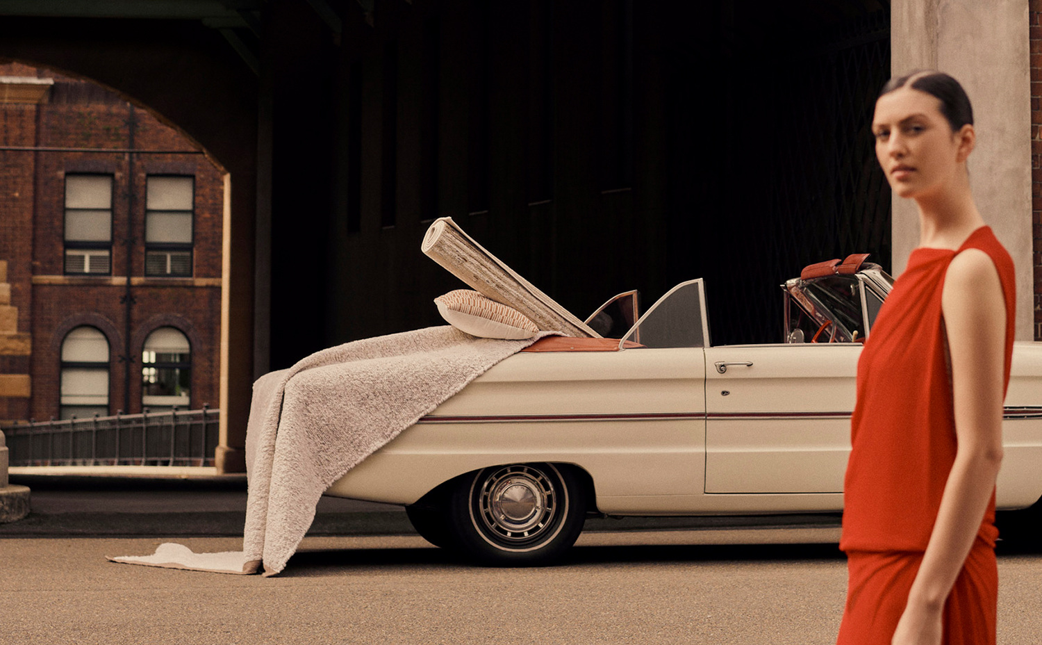 Fashion shot of a woman in a red dress next to a vintage car with a rolled-up carpet and cushion, set against a brick building backdrop.