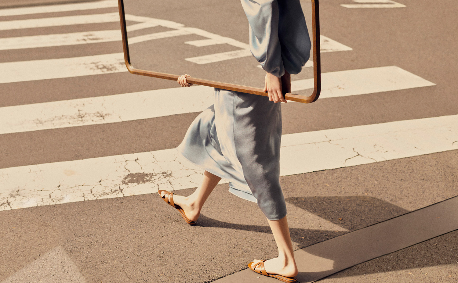 Woman in a blue dress walks in a street holding a square wooden frame on a sunny day, showcasing fashion and street photography.