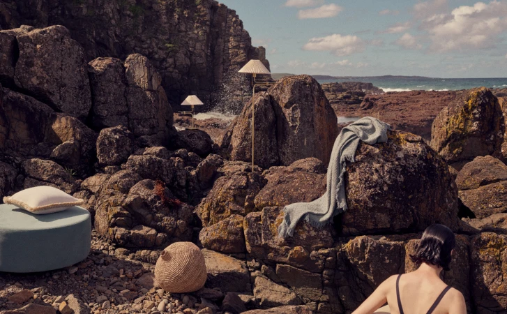 Coastal living scene with lamps, cushions, and a blanket arranged on rugged rocks overlooking the ocean on a sunny day with blue skies.
