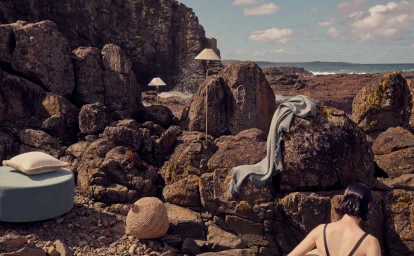 Woman with wet dark hair viewed from behind sitting among rugged dark rocks outdoors by the ocean with home decor items