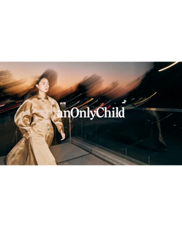 Model wearing a gold satin dress walking past a railing at dusk with motion blurred light streaks in the background.