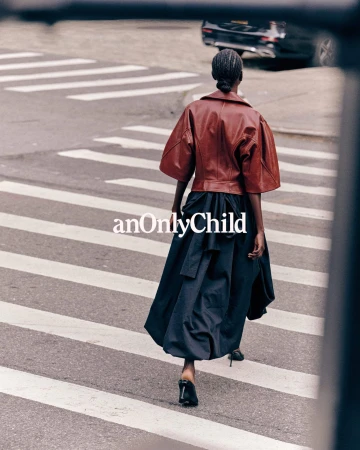 Model wearing a brown leather jacket and dark skirt walking across a city crosswalk
