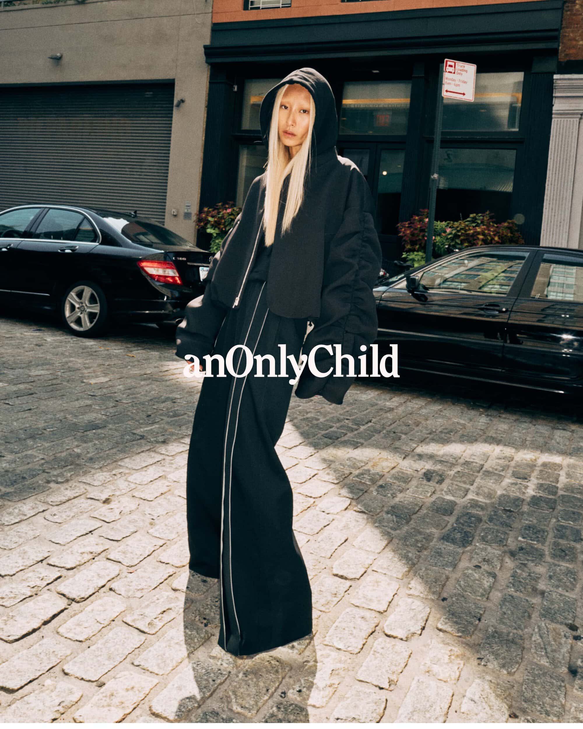 Model in black hoodie and wide pants standing on a cobblestone street with parked cars for anOnlyChild