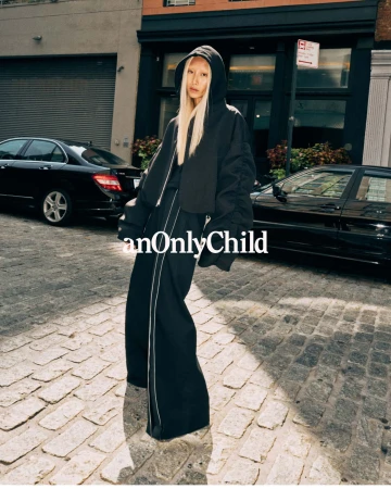 Model in black hoodie and wide pants standing on a cobblestone street with parked cars for anOnlyChild