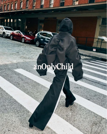 Figure in dark hooded sweatshirt and trousers walking across a city crosswalk with parked cars and brick building in the background