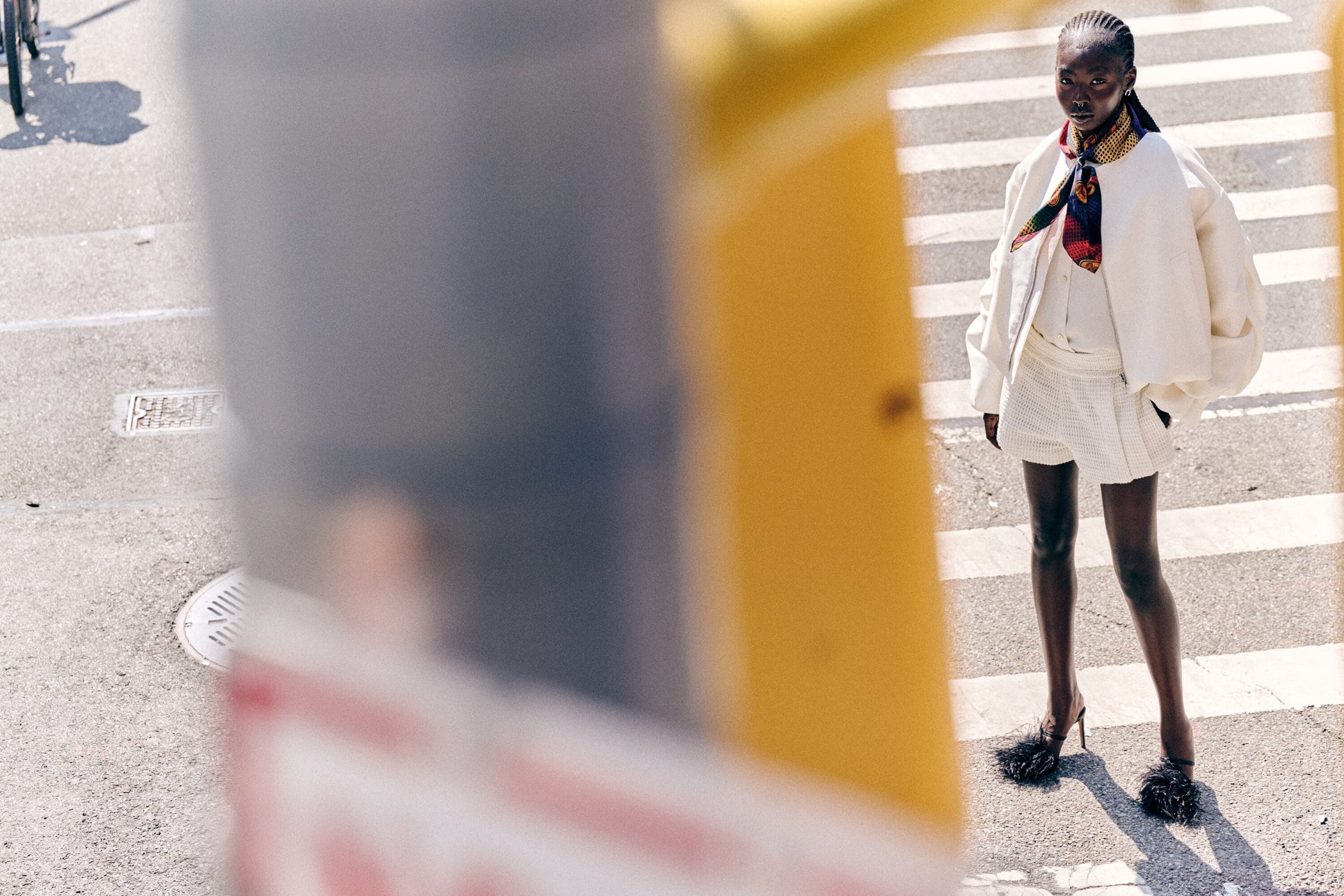 Model standing on a pedestrian crosswalk wearing a white jacket, shorts, patterned neck scarf, and feathered heels, framed through a street pole