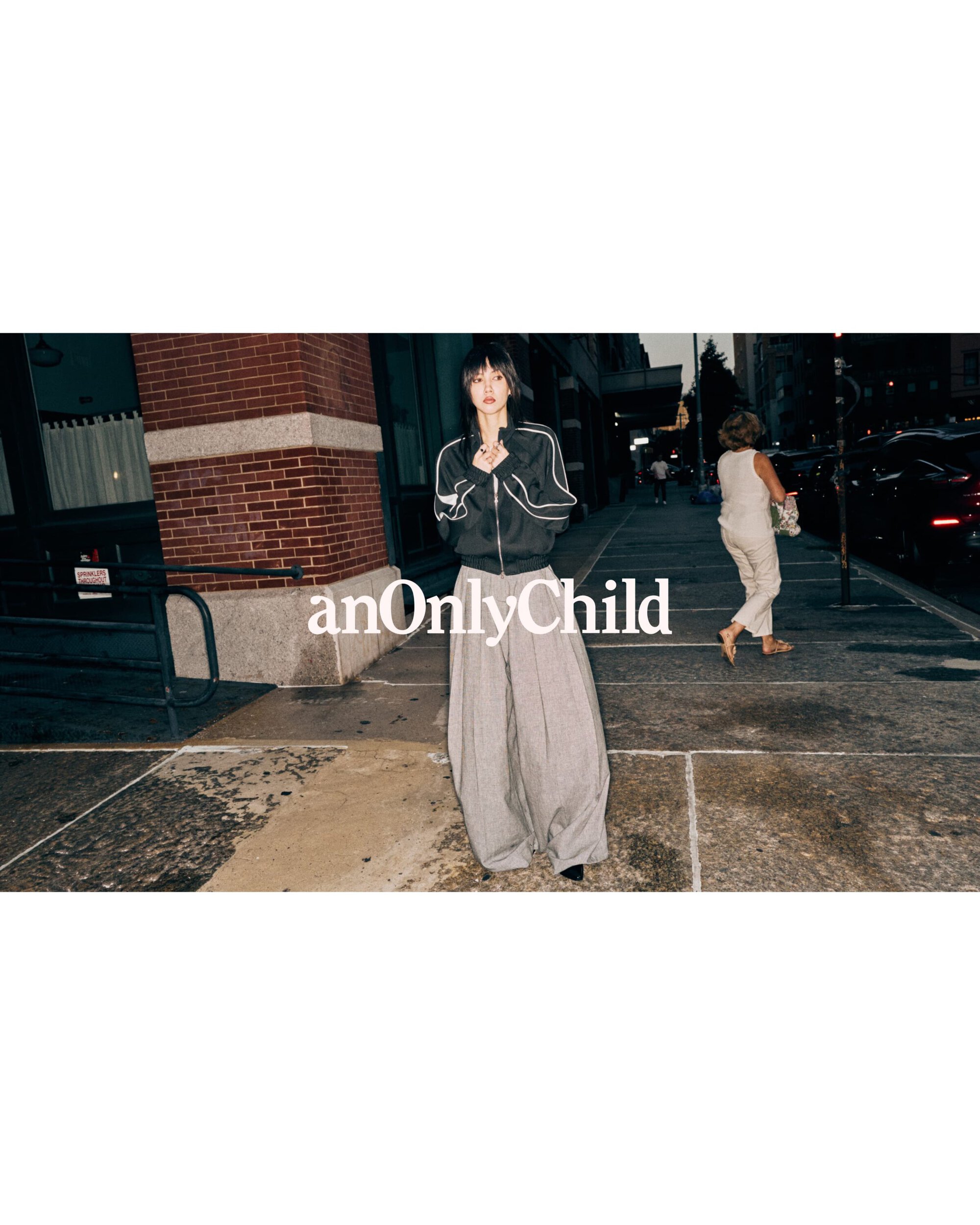 Model wearing dark zip-up jacket and wide grey trousers standing on a city sidewalk for anOnlyChild Spring 2025 Ad Campaign.