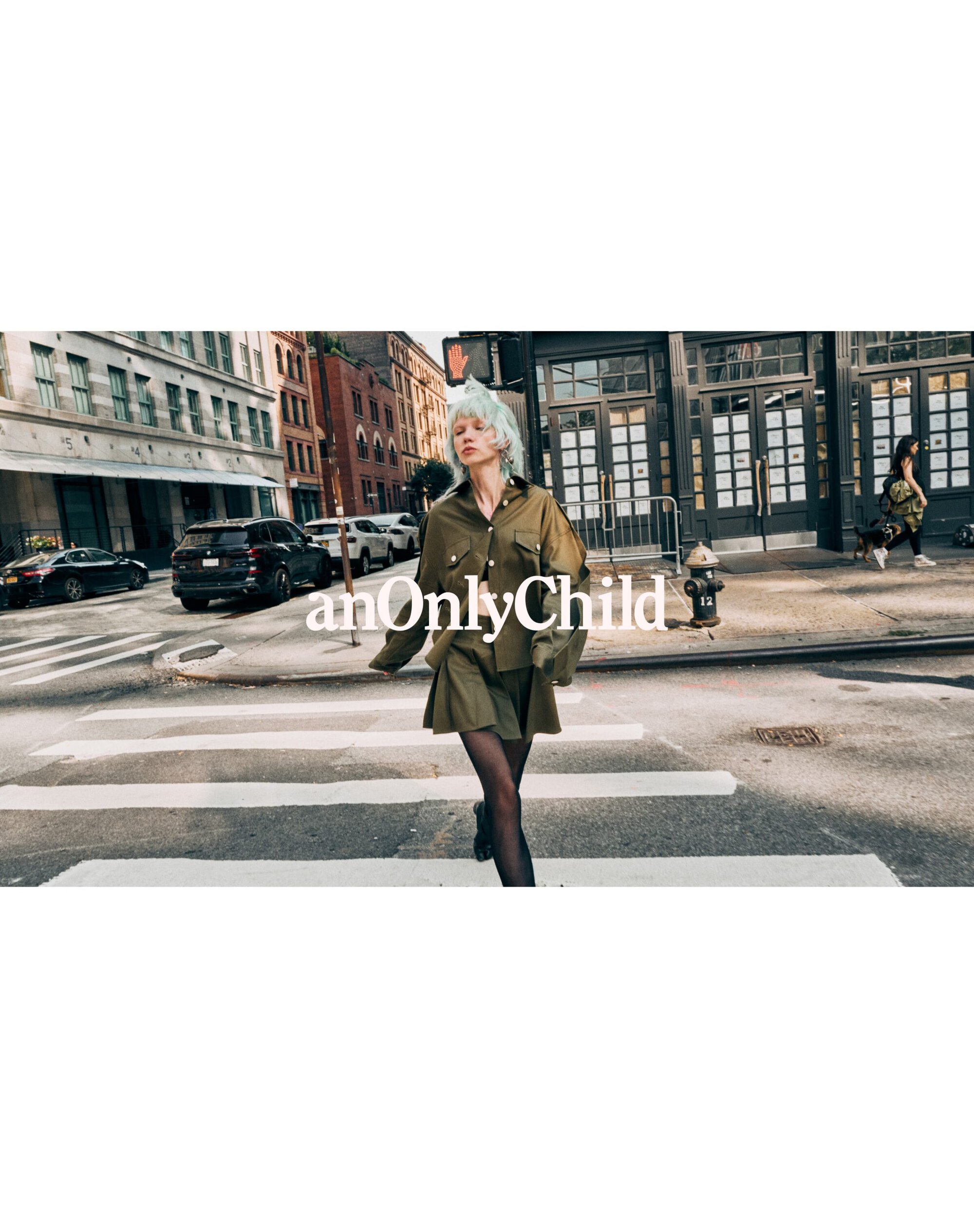 Model with light blue hair wearing olive green shirt and skirt walking across a city crosswalk for anOnlyChild
