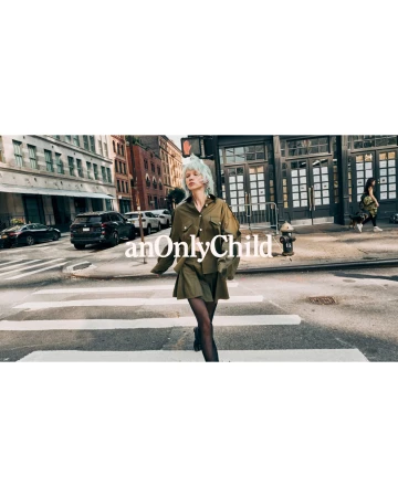 Model with light blue hair wearing olive green shirt and skirt walking across a city crosswalk for anOnlyChild