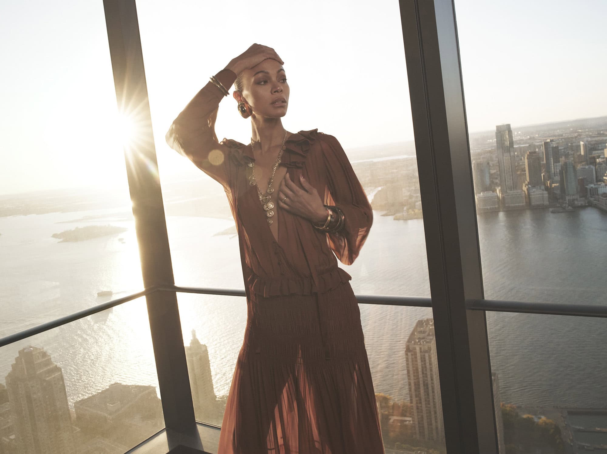 Zoe Saldaña in a sheer brown dress standing by a high-rise window overlooking a city skyline at sunset