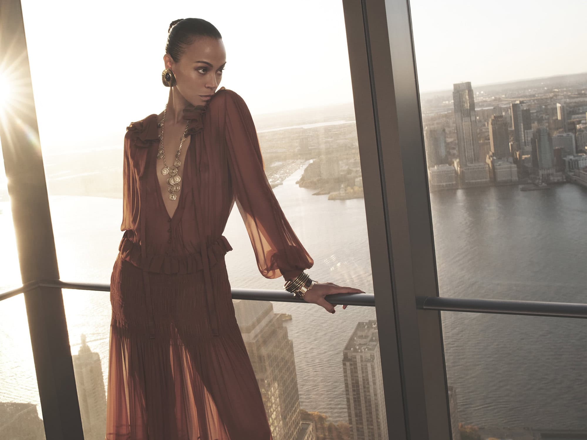 Zoe Saldana posing in a sheer brown blouse and skirt against a high-rise city view for Saint Laurent Spring 2025 campaign.