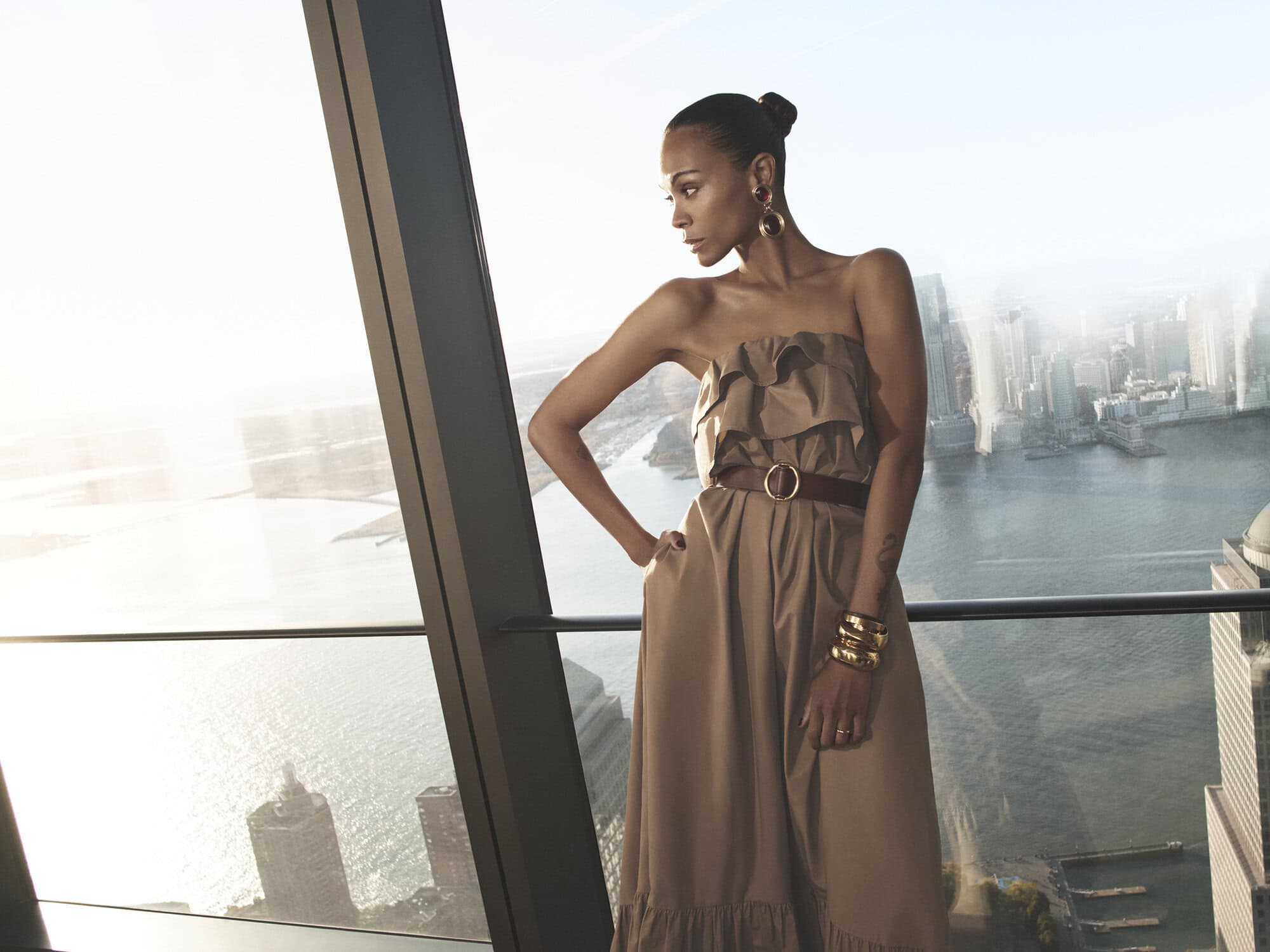 Zoe Saldaña wearing a brown strapless dress and gold jewelry by a floor-to-ceiling window overlooking a city skyline and water.