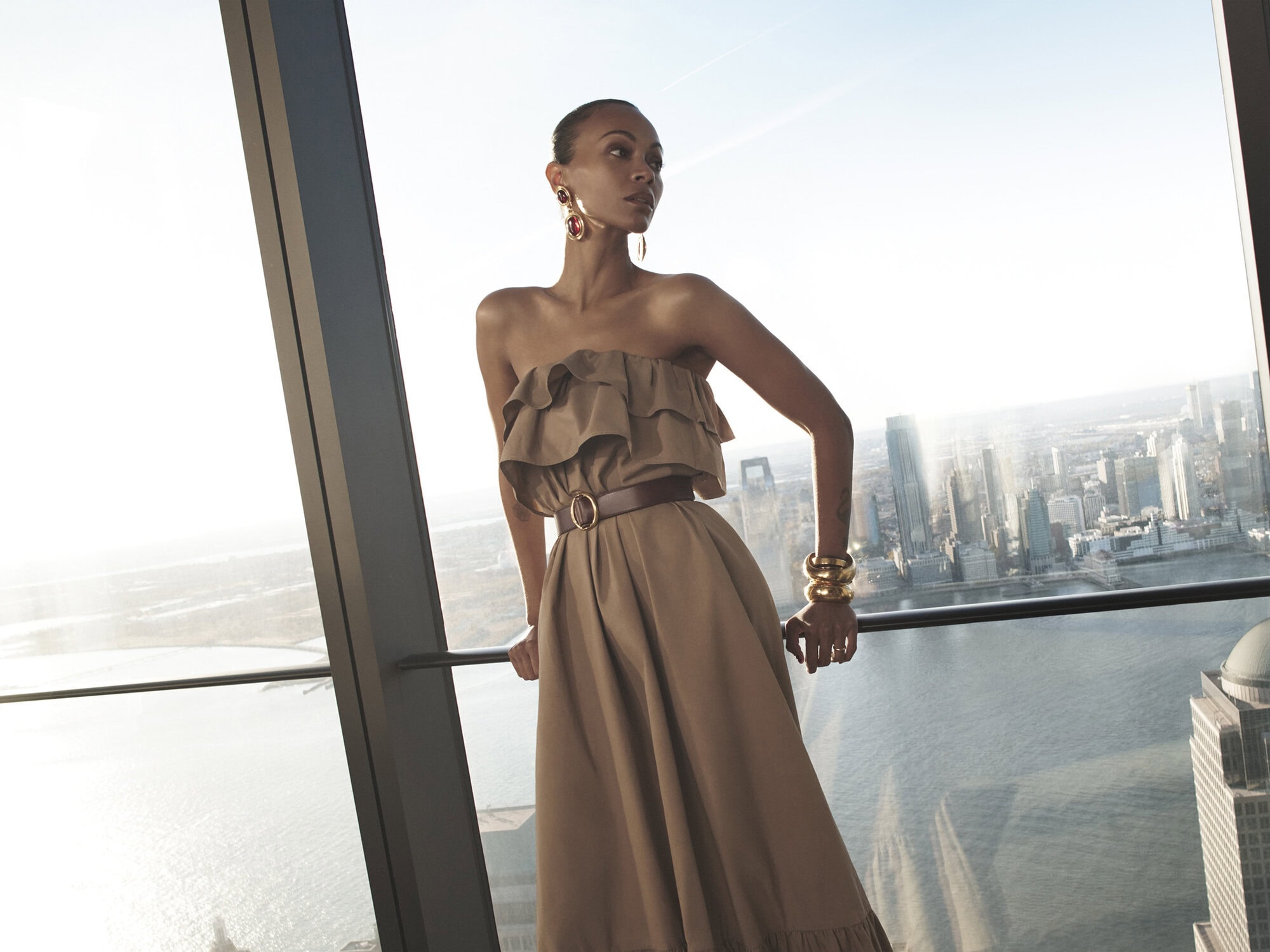 Zoe Saldaña wearing a strapless tan dress and statement jewelry leaning against a skyscraper window overlooking a city skyline