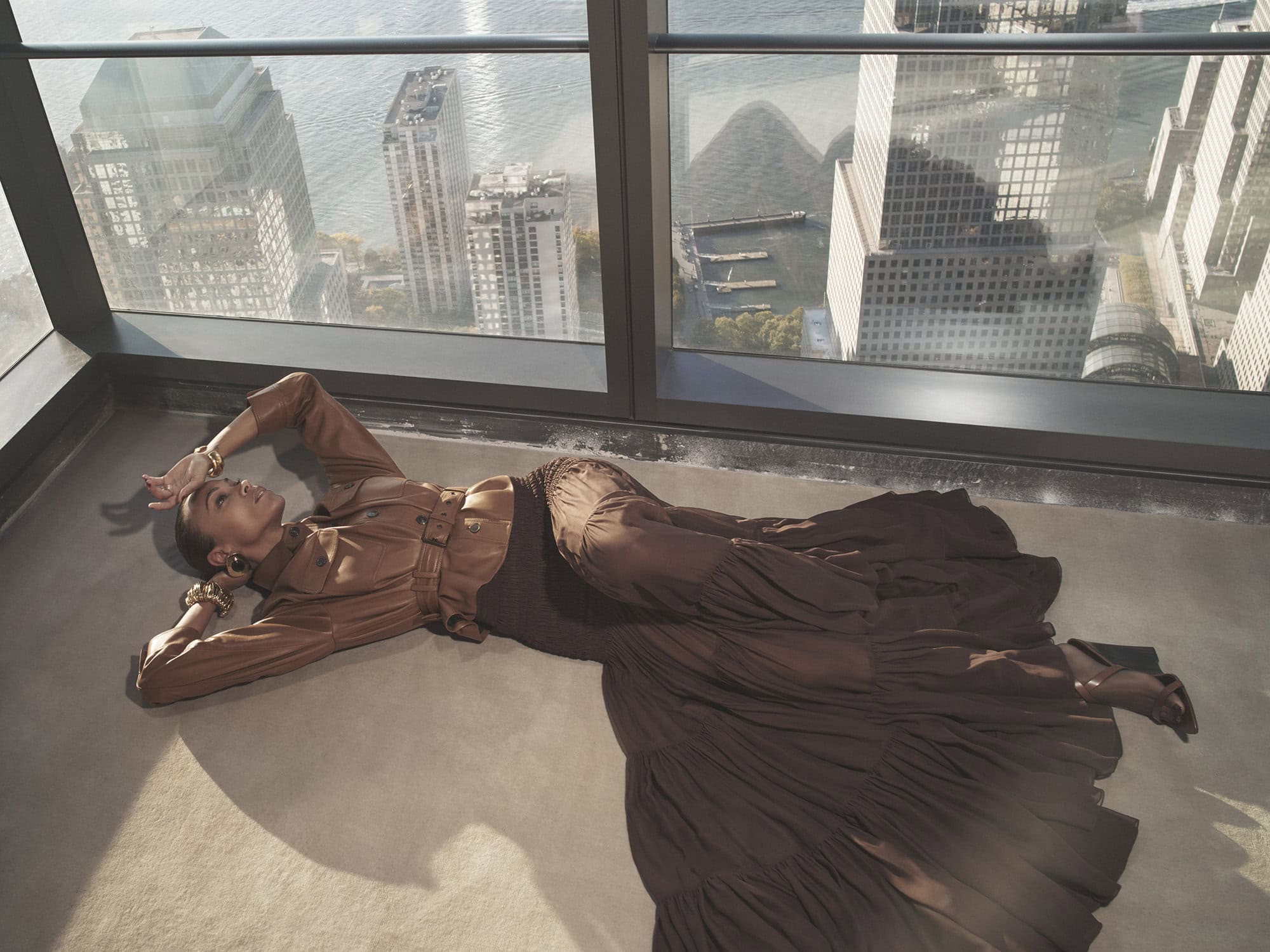 Woman in brown belted leather jacket and long ruffled skirt lying on floor against large skyscraper windows