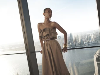 Zoe Saldaña wearing a strapless tan dress and statement jewelry leaning against a skyscraper window overlooking a city skyline