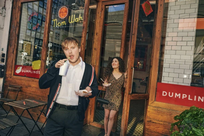 Man drinking from a cup outside a storefront while a woman stands in the doorway