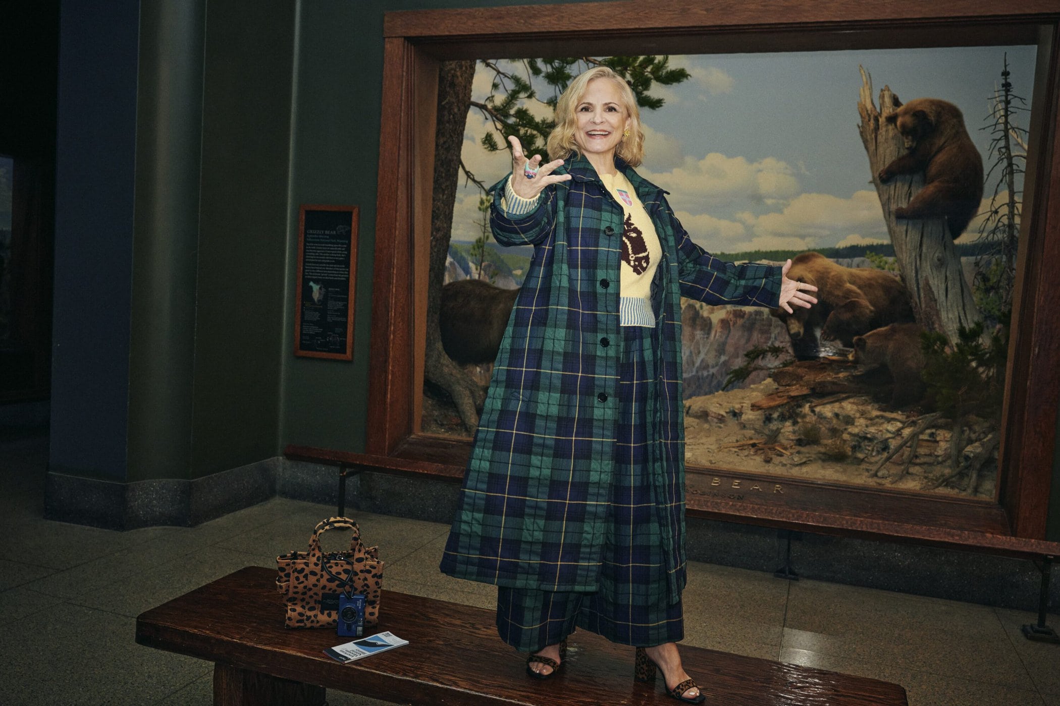 Amy Sedaris wearing a green plaid coat standing in a museum exhibit with a large bear diorama backdrop