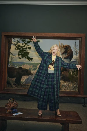 Amy Sedaris wearing a plaid coat standing on a wooden bench in a museum diorama for Nordstrom