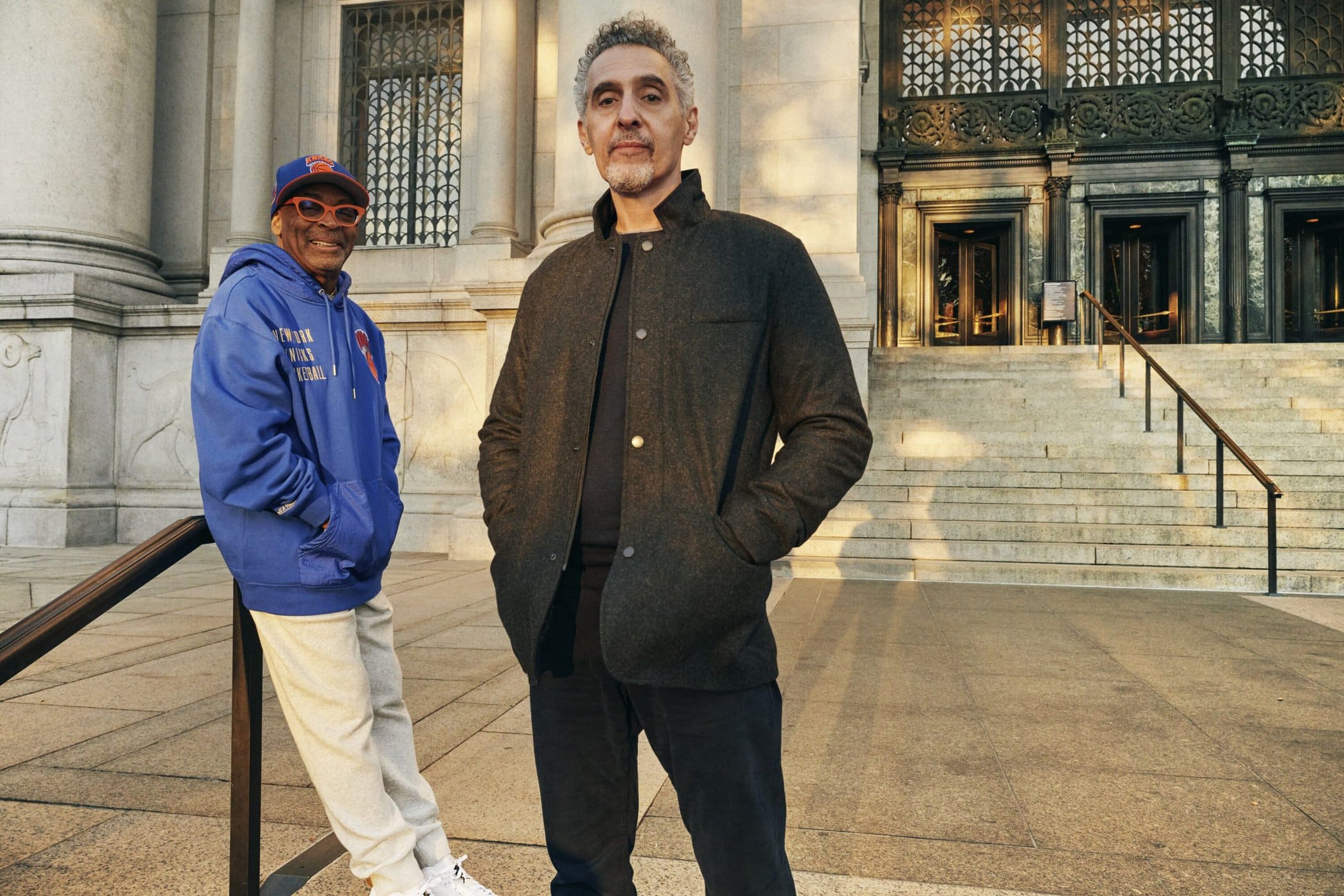 Spike Lee in a blue hoodie and John Turturro in a dark jacket pose outside a stone building in New York City.