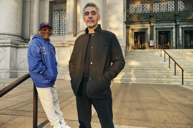 Spike Lee in a blue hoodie and John Turturro in a dark jacket pose outside a stone building in New York City.