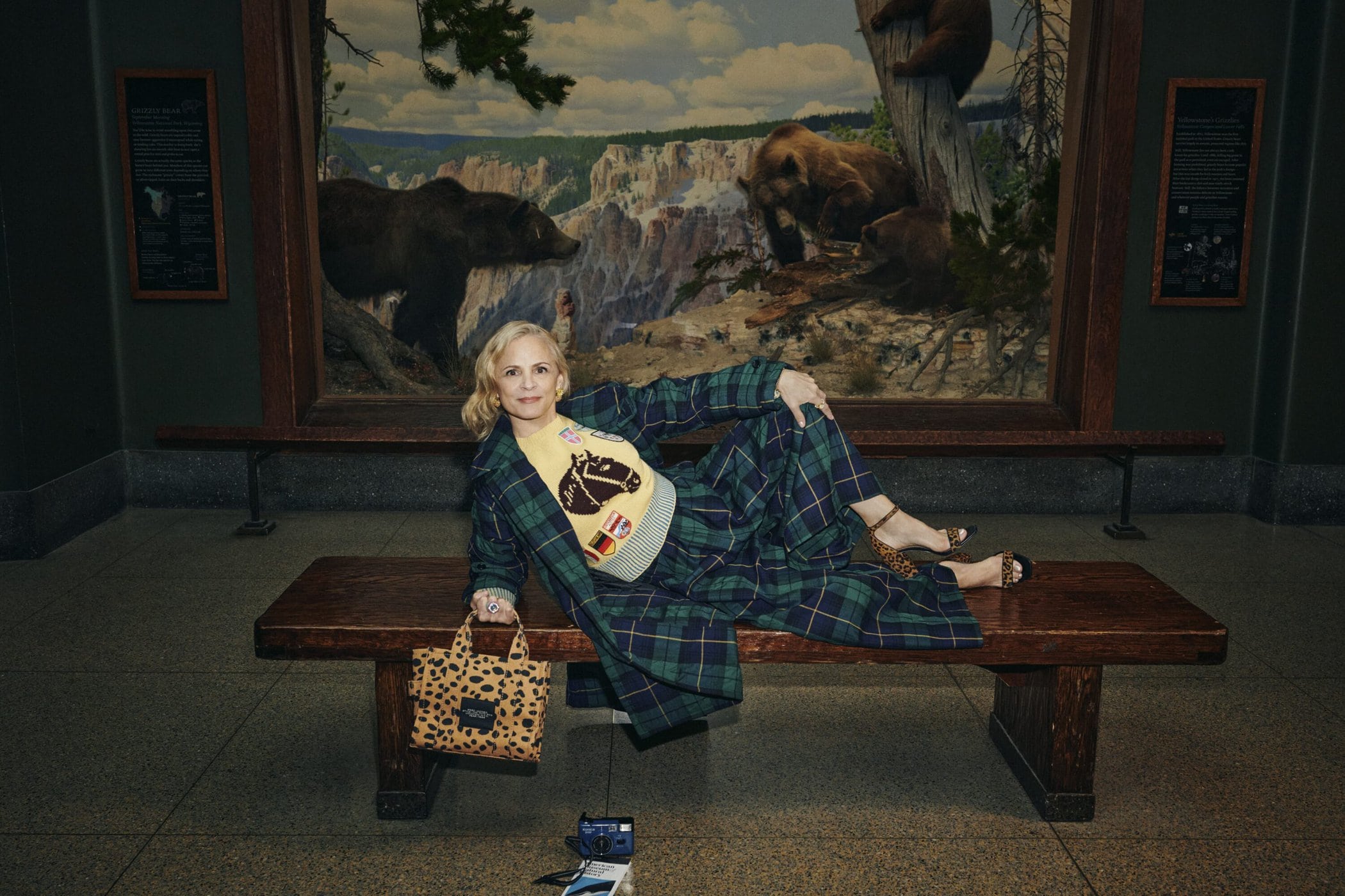 Woman in plaid coat and matching trousers lying on a wooden bench inside a museum with a bear diorama background.